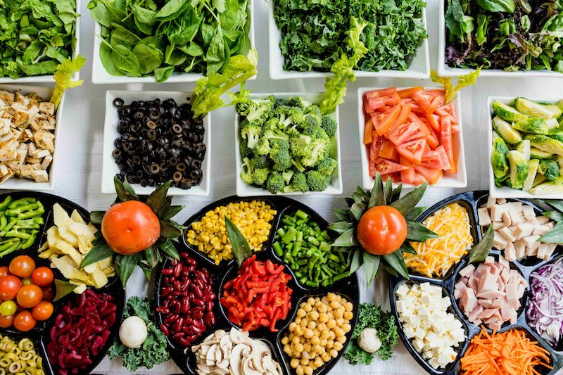 Salad bar trays of spinach, kale, olives, broccoli, tomatoes, corn, beans, peppers and chickpeas seen from above
